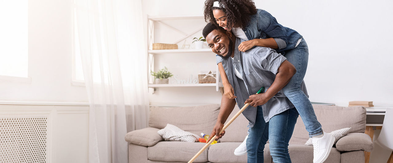Pareja divirtiéndose mientras trapea el piso de la sala con productos de limpieza, fomentando la limpieza del hogar compartida y el bienestar.